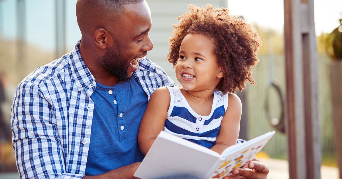 Father reading a board book with his toddler daughter outdoors, smiling and bonding during story time